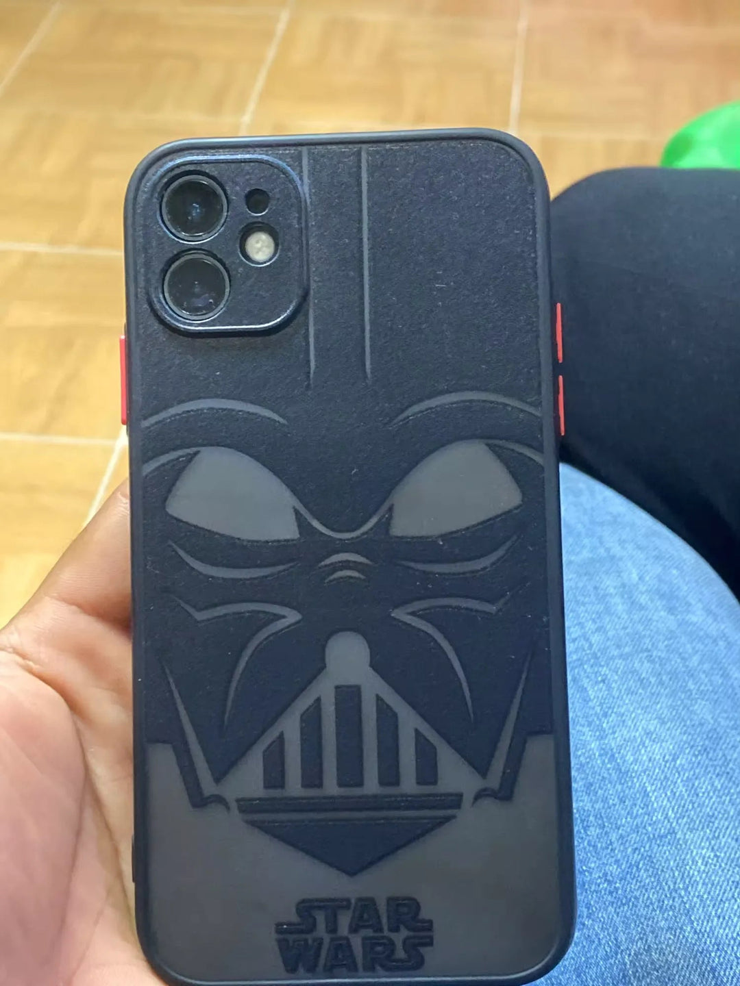 A person is holding the Vader Mask for iPhone, an anti-scratch smartphone case featuring a design of Darth Vader’s iconic face. The background includes a tiled floor and part of someone’s leg wearing jeans.