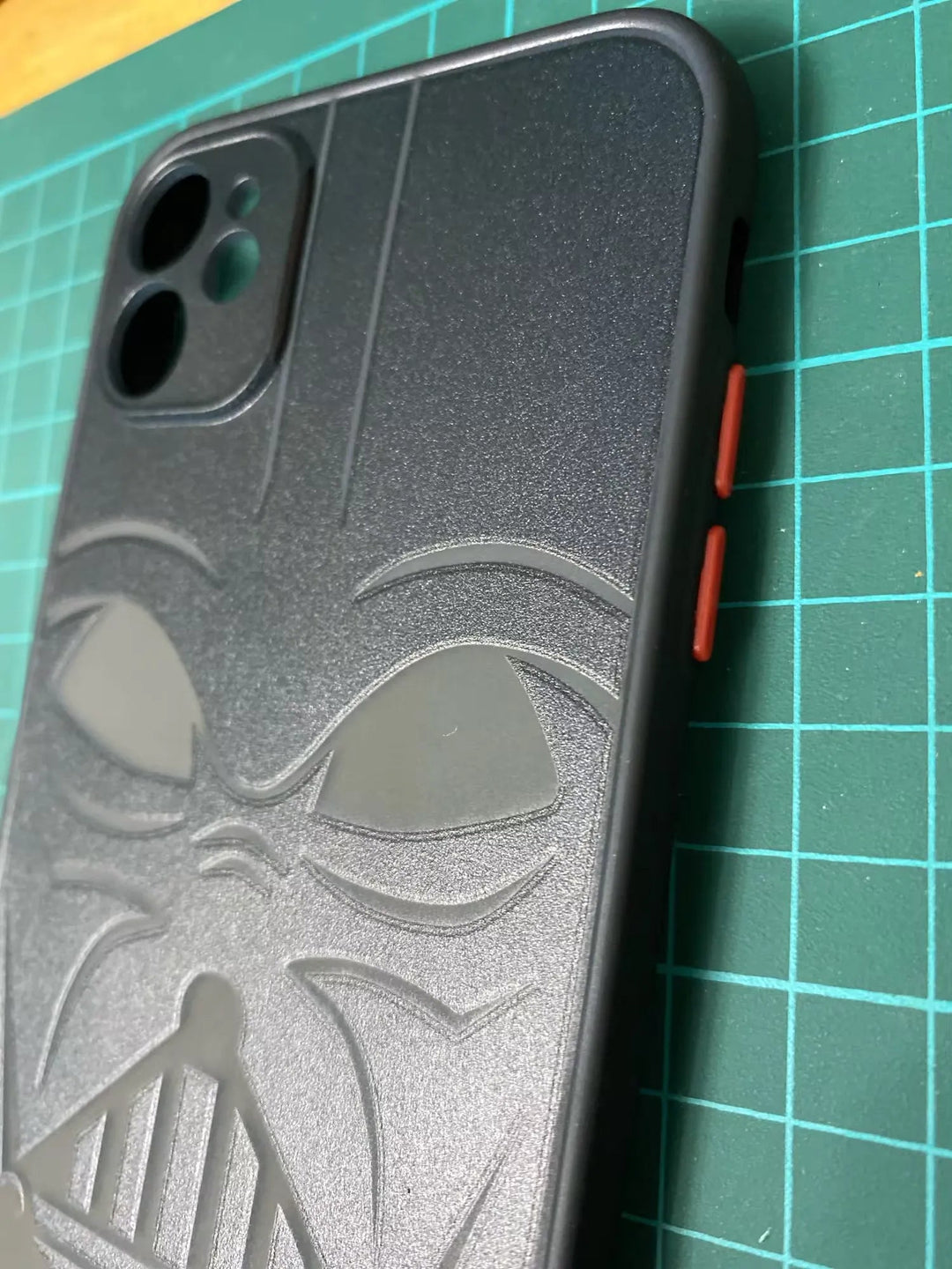 A black anti-scratch smartphone case from the Vader Mask for iPhone collection is shown, featuring a Darth Vader mask design and red side buttons. The case is placed on a green cutting mat with grid lines.