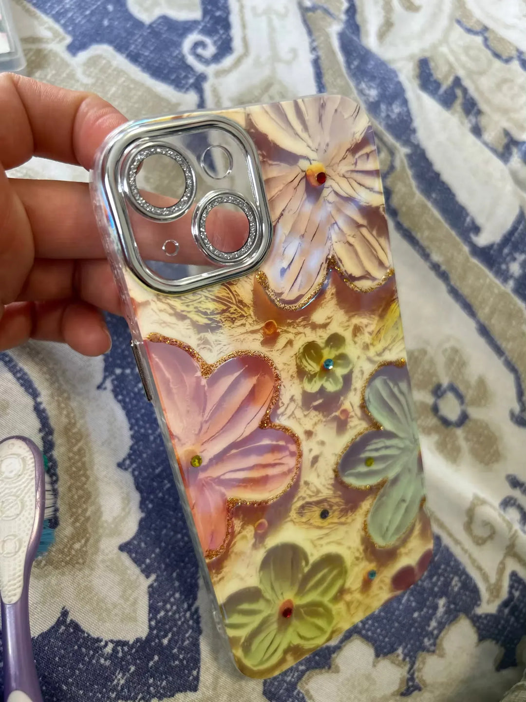 A close-up photo of a clear phone case with a 3D floral design.
 The flowers are painted in pastel colors and adorned with glitter and rhinestones. The case is being held in someone's hand.